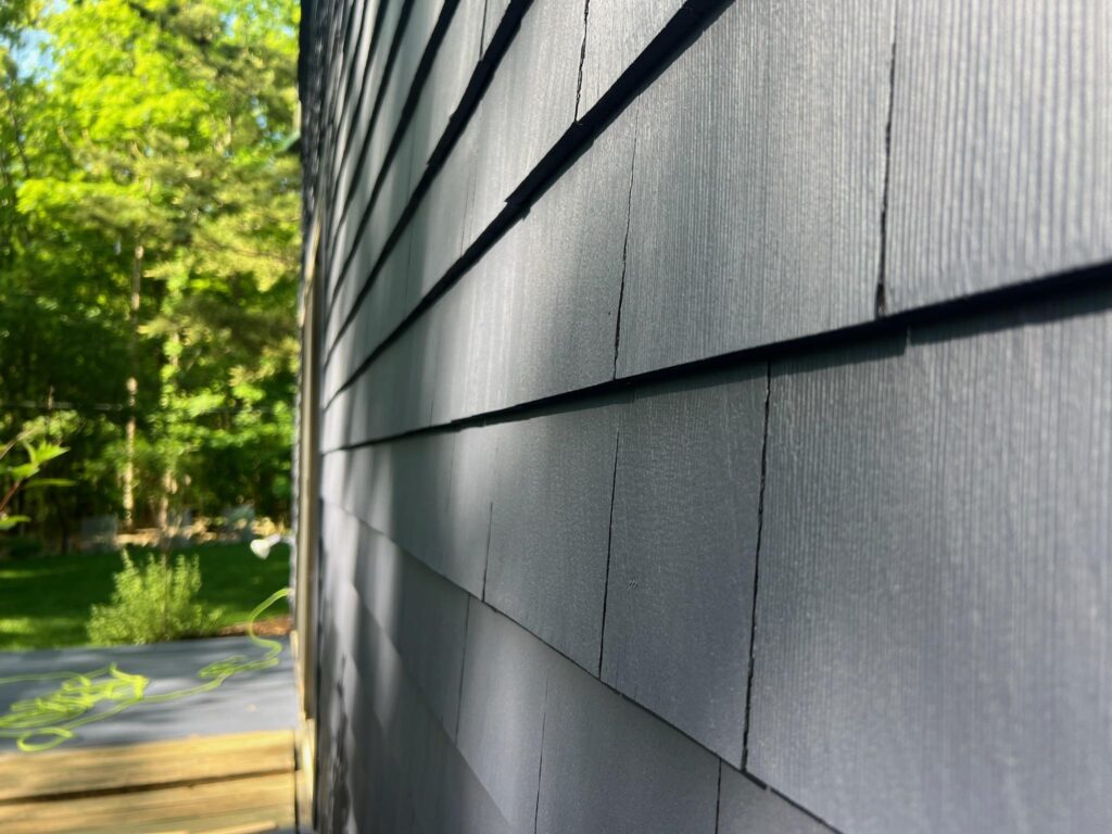 Close-up of gray exterior painting shingled siding on a sunlit house, with shadows creating a textured pattern. Green trees and a garden are visible in the background.