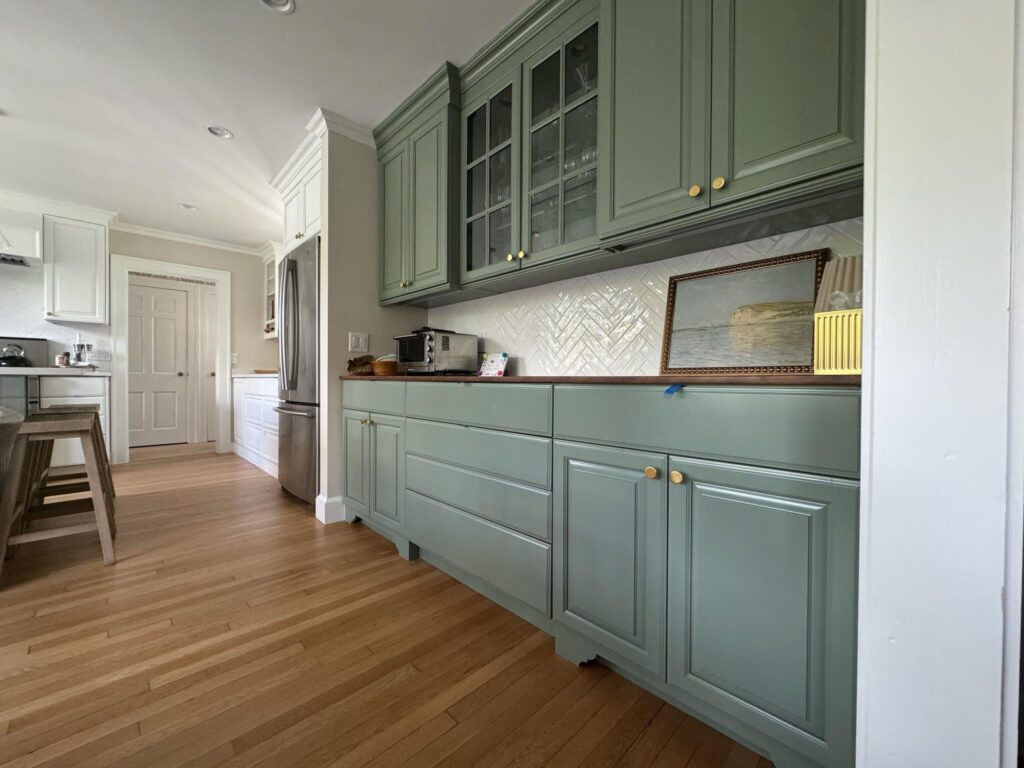 Modern kitchen with light green cabinets and wooden floor. Stainless steel fridge, white herringbone backsplash, and a framed painting add elegance.