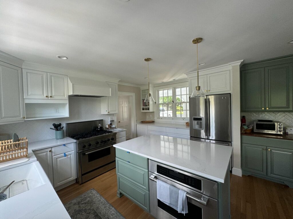 Modern kitchen with white and green cabinetry, stainless steel appliances, and a large island. Bright natural light streams through a window, creating an inviting atmosphere.