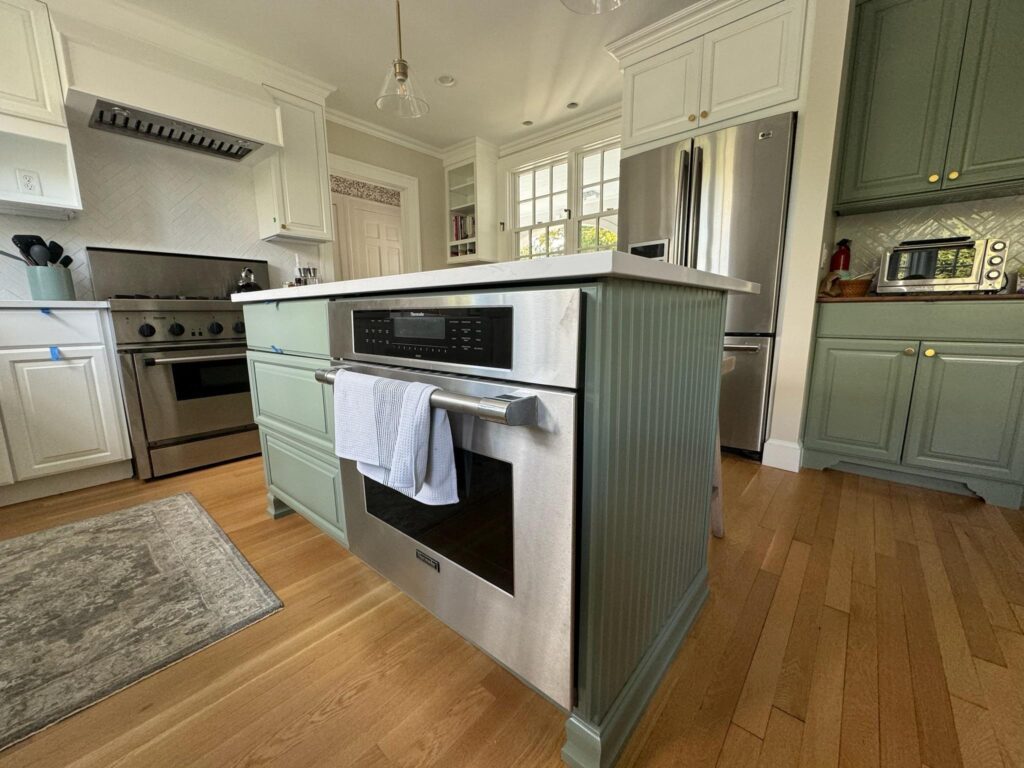 Cabinet refacing with light green island featuring a built-in oven and towel. Stainless steel appliances, white cabinetry, and wooden floors create a cozy atmosphere.