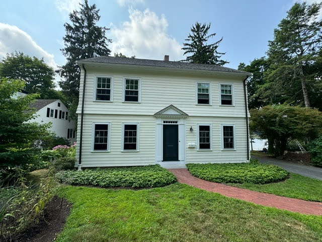 Two-story white house with black shutters and a central door, surrounded by trimmed bushes and trees. A red brick path leads to the entrance.
