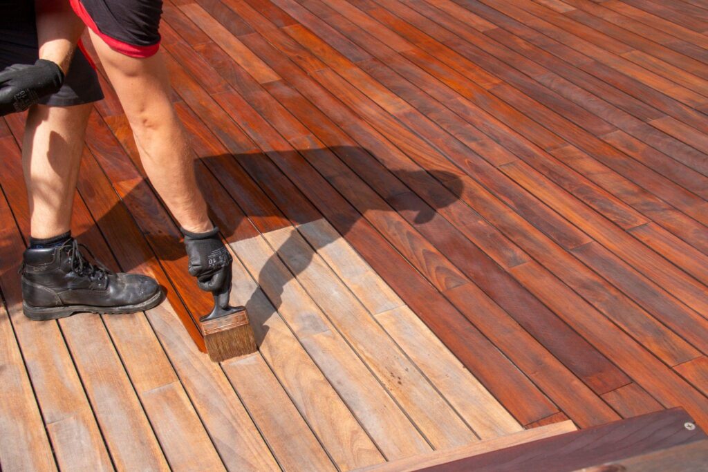 Professional House Painter applying stain to a wooden deck with a brush. The wood has rich, warm tones, and the person wears work boots and gloves.